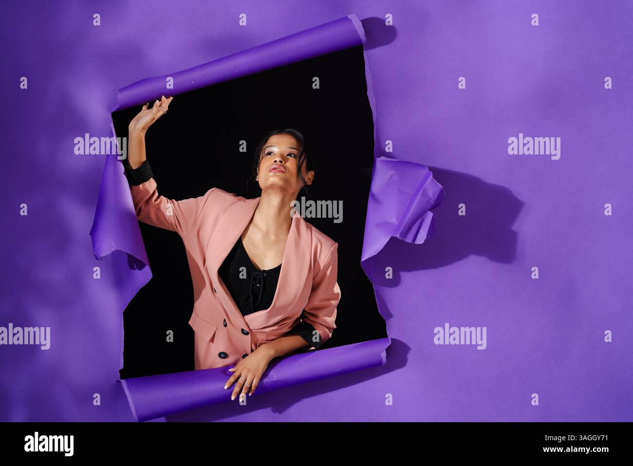 Sie bricht durch die lila Papierwand, eine Frau in rosa Blazer, die Freiheit ausdrückt. Stärke, Ermächtigung, Ausdruck, Befreiung, Selbstvertrauen Stockfoto