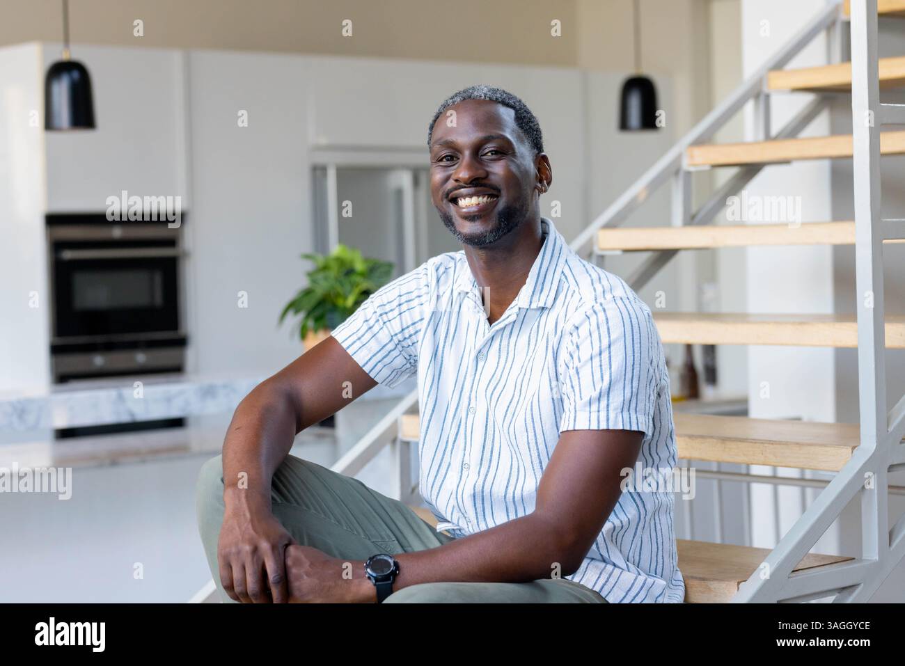 Lächelnder afroamerikanischer Mann, der auf einer Treppe in einem modernen Haus sitzt und einen entspannten Morgen genießt. Lifestyle, Entspannung, lässig, Glück, Innenraum, Contem Stockfoto