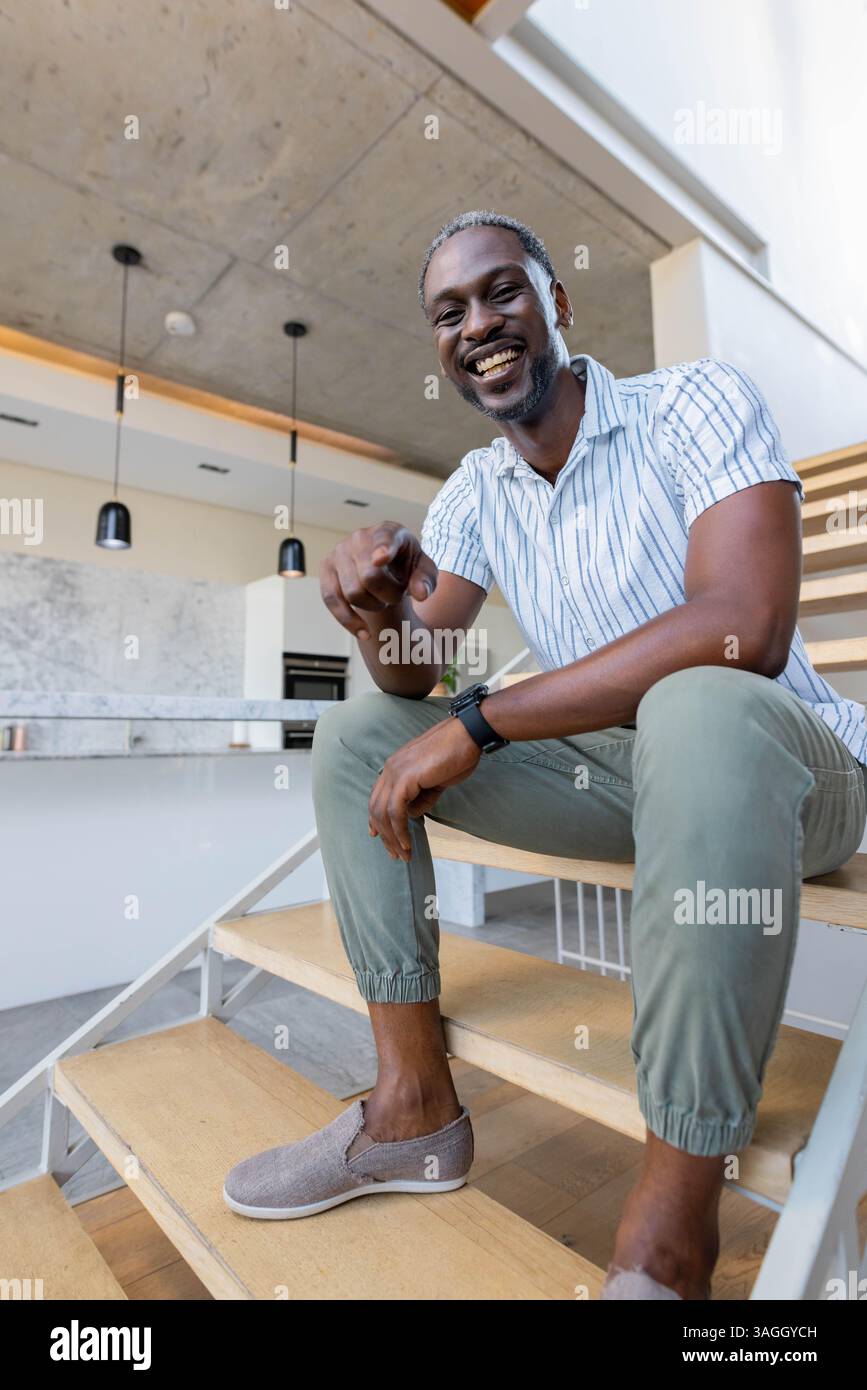 Ein lächelnder Afroamerikaner sitzt zu Hause auf der Treppe und zeigt spielerisch auf die Kamera. Glücklich, lässig, Lifestyle, Indoor, männlich, Treppenaufgang Stockfoto