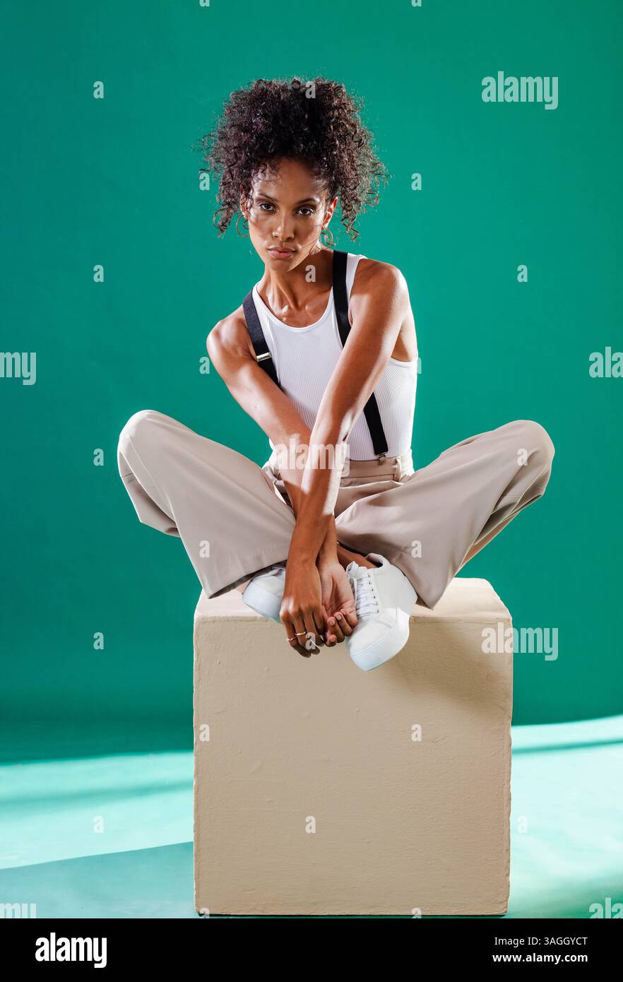 Auf Cube sitzend, eine Frau mit Hosenträgern und Sneakern in selbstbewusster Haltung. Mode, Selbstvertrauen, Style, Urban, trendy, Schuhe Stockfoto