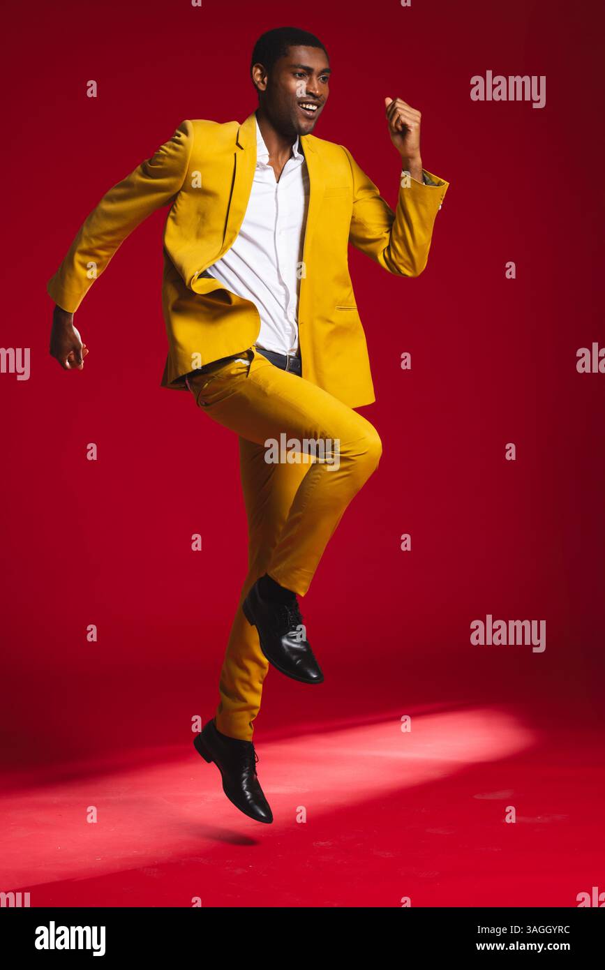Mann in leuchtendem gelbem Anzug, der fröhlich vor rotem Studio-Hintergrund springt. Mode, bunt, Enthusiasmus, Energie, Studio-Aufnahme, dynamisch Stockfoto