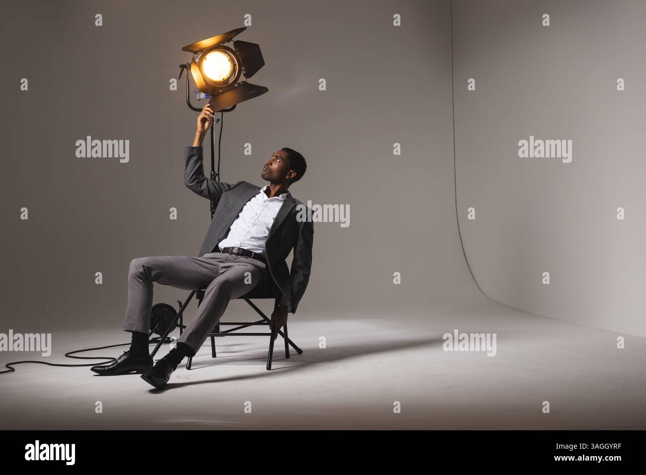 Stilvoller Mann im Anzug, der das Studio-Licht anpasst, während er auf dem Stuhl sitzt. Fotografie, Beleuchtung, Porträt, professionell, Mode, Kreativität Stockfoto