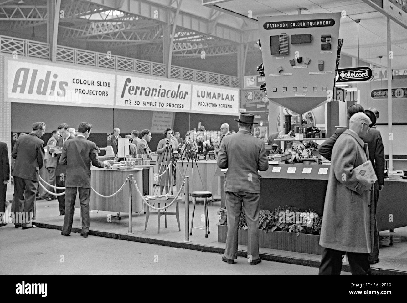 Besucher einer Fachmesse für Film- und Fotohersteller und Zulieferer in Olympia, West Kensington, Hammersmith, London, England, UK in c.1960. Am Paterson-Stand waren Dunkelkammer und Fotoausrüstung zu sehen. Dahinter befinden sich Banner für Aldis Diaprojektoren, das italienische Unternehmen Ferrania, das für ihren Ferraniacolor-Fotofilm und Lumaplak-Projektionsleinwände wirbt. Das ist von einem alten Schwarzweiß-negativ eines Amateurfotografen – ein Vintage-Foto aus den 1950er/60er Jahren. Stockfoto