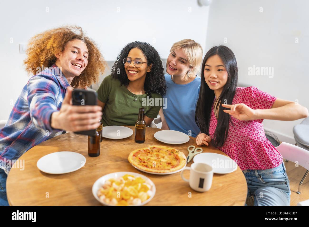 Vier verschiedene Freunde machen ein Selfie mit einem Smartphone, während sie zu Hause ein Mittagessen mit Pizza und Bier genießen Stockfoto