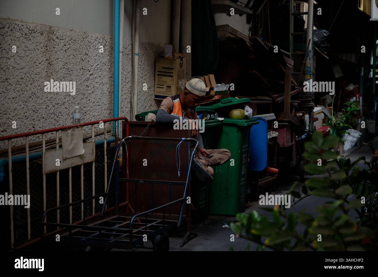 Der Müllsammler macht eine Pause neben seinem Wagen und den Mülltonnen in einer versteckten Bangkok Gasse Stockfoto