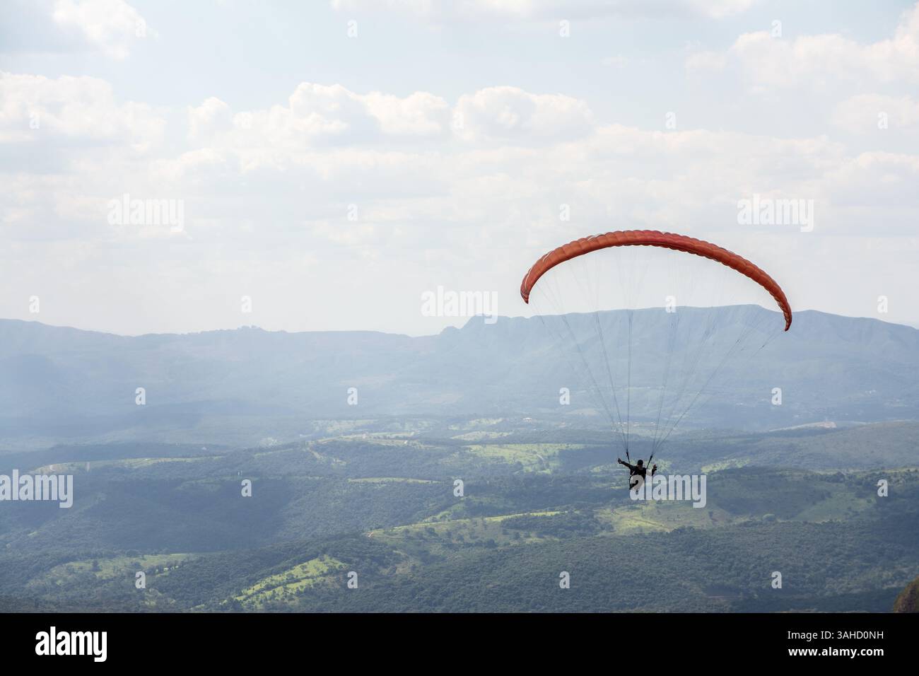 Einsame Gleitschirmflieger gleiten durch die Luft über eine grüne, bergige Kulisse und teilweise bewölkten Himmel. Stockfoto