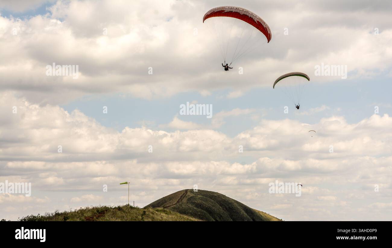 Zwei Gleitschirmflieger gleiten durch die Luft über eine grüne, bergige Kulisse und teilweise bewölkten Himmel. Stockfoto