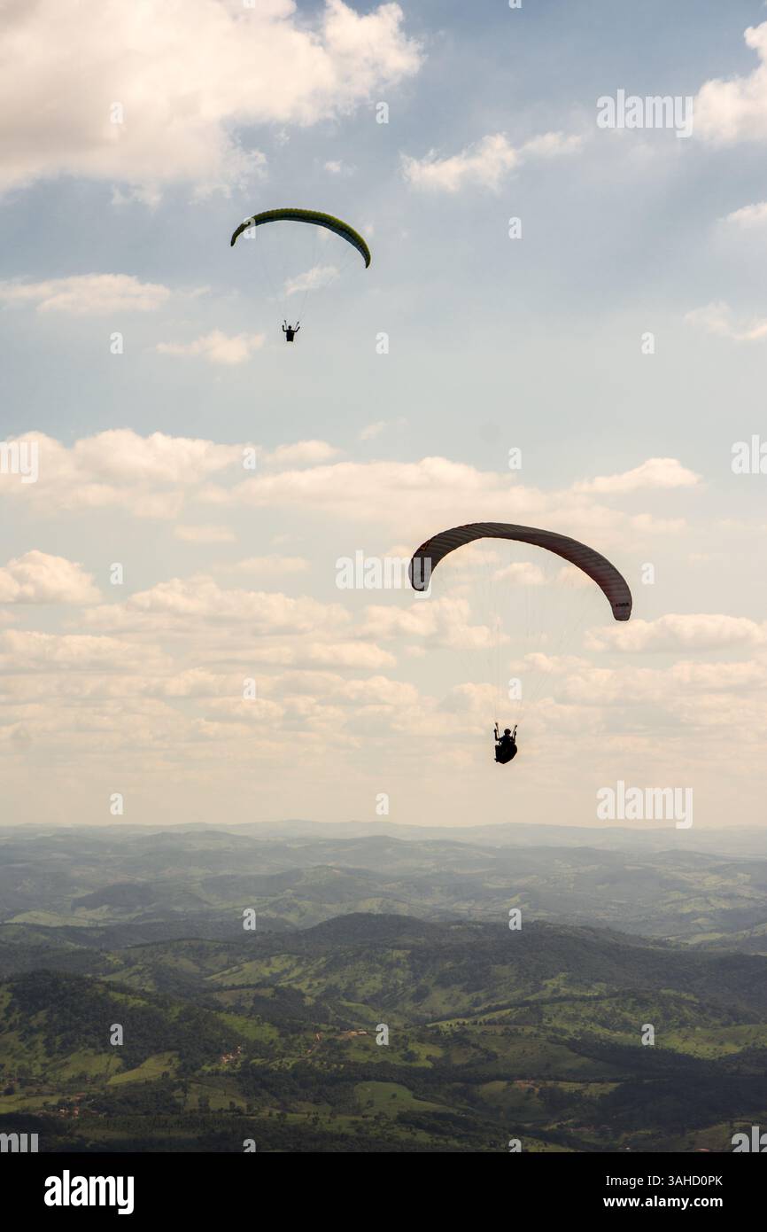 Zwei Gleitschirmflieger gleiten durch die Luft über eine grüne, bergige Kulisse und teilweise bewölkten Himmel. Stockfoto