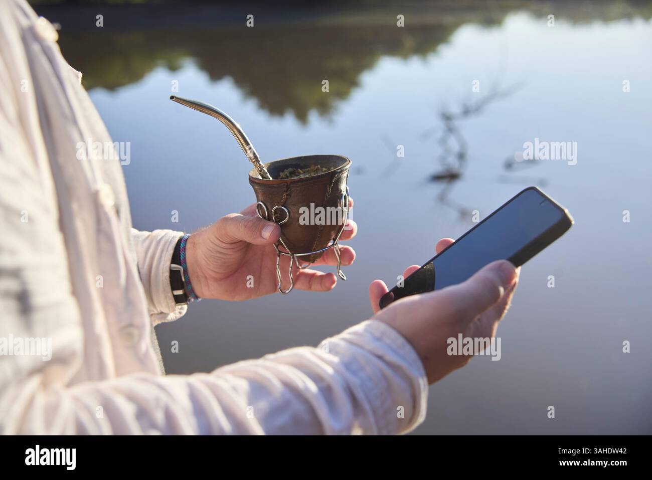 Person, die sein Smartphone benutzt, während er draußen Mate trinkt. Selektiver Fokus auf die Hand mit dem Infusion, traditionelles Getränk der Wahl für Argentinien Stockfoto