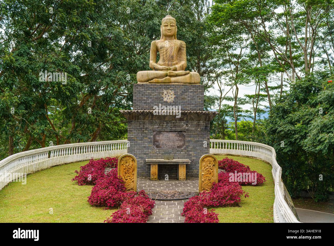 Eine ruhige Buddha-Statue befindet sich auf einem Backsteinsockel, umgeben von üppigem Grün und lebendigen Blumen in Gampola, Sri Lanka, und symbolisiert Frieden und Geist Stockfoto
