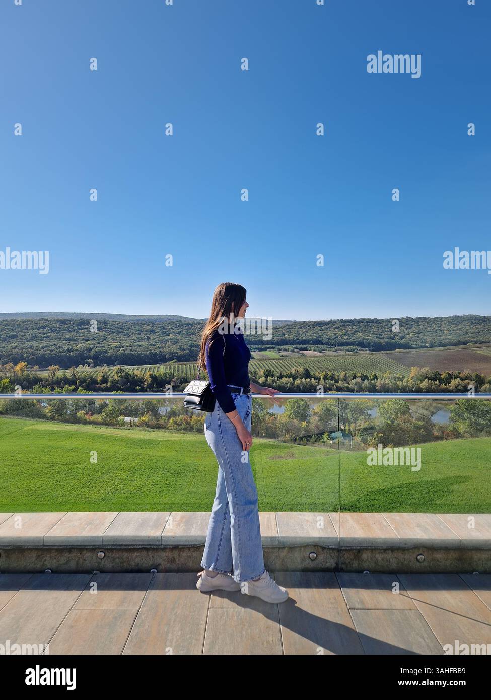 Die junge Frau steht auf einer Terrasse mit einem Glasgeländer mit Blick auf eine weite, grüne Landschaft mit sanften Hügeln unter einem klaren blauen Himmel Stockfoto