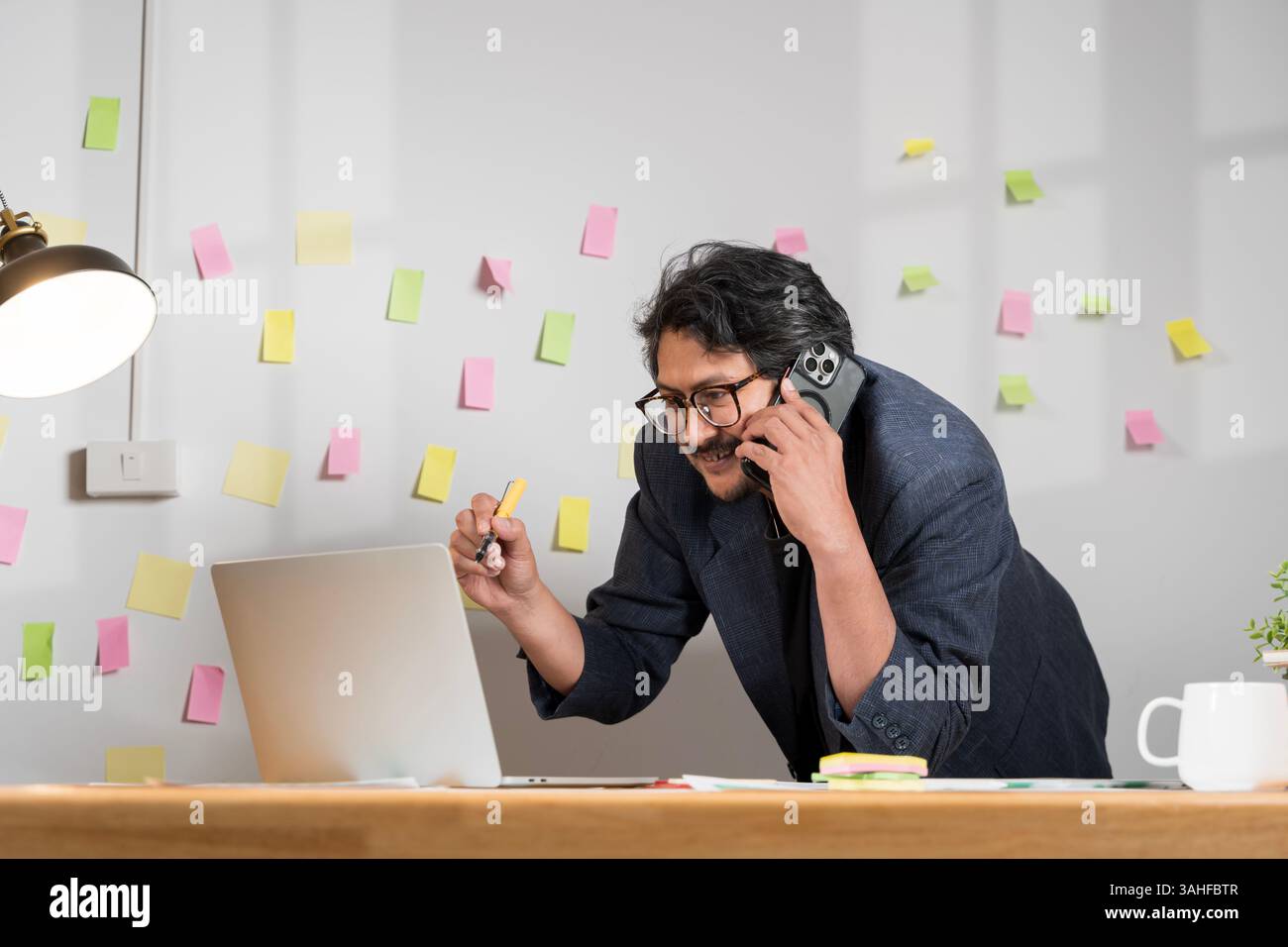 Ein mittelgroßer kaukasischer Mann in Geschäftskleidung arbeitet intensiv an einem Laptop, während er ein Smartphone handhabt, und reflektiert Multitasking in einem eleganten Firmenstil Stockfoto