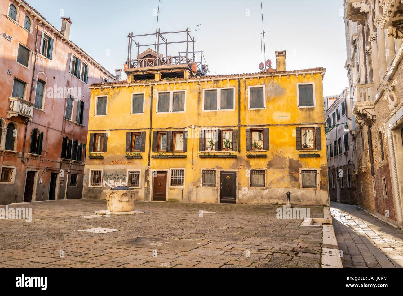 Gelbes Gebäude mit verwitterter Fassade, kleinem Platz und Brunnen unter klarem blauem Himmel. Venedig, Italien Stockfoto