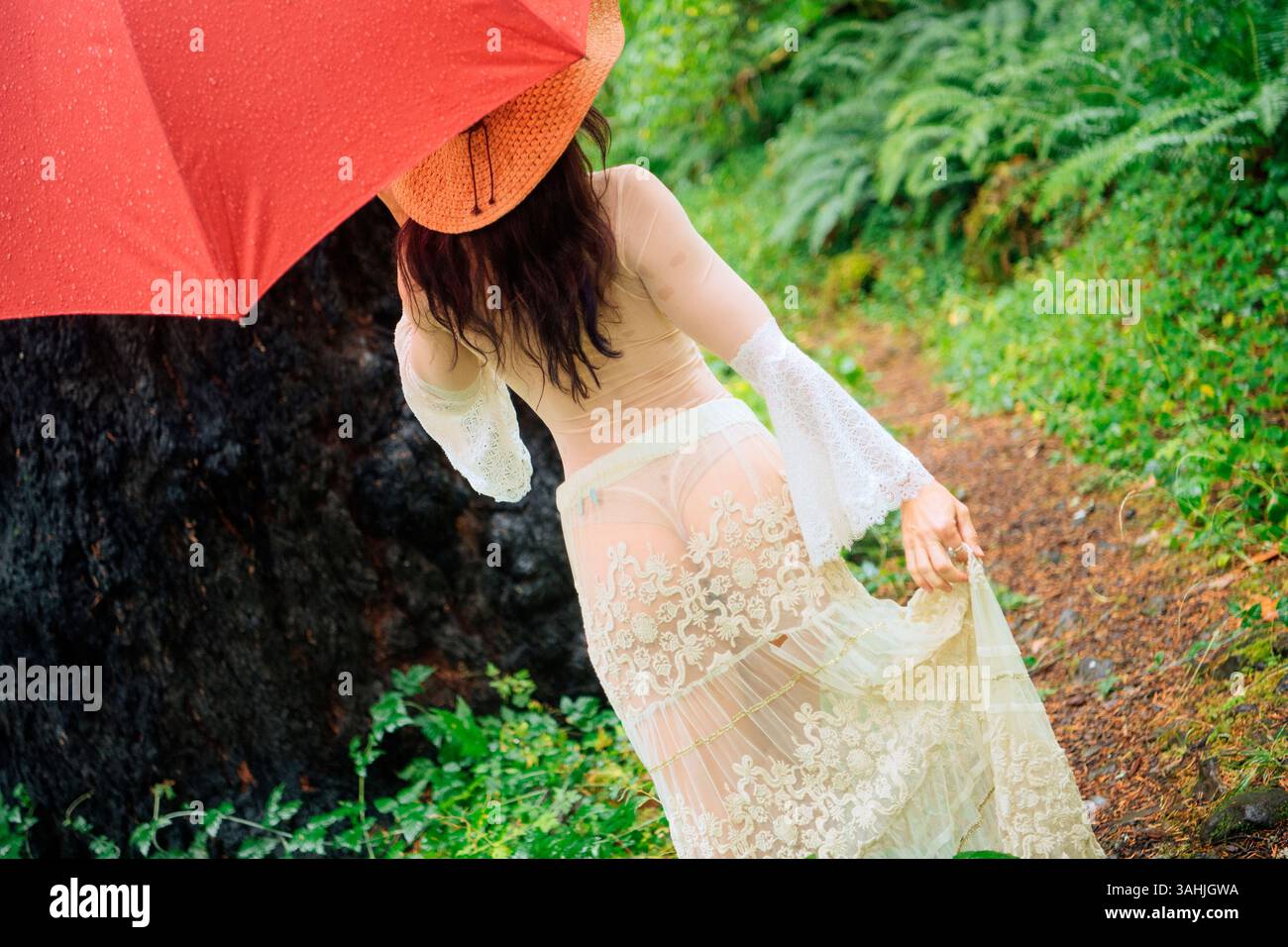 Frau in einem transparenten Kleid mit einem roten Regenschirm in einer üppig grünen Waldlandschaft. Silver Falls State Park, Oregon, USA Stockfoto