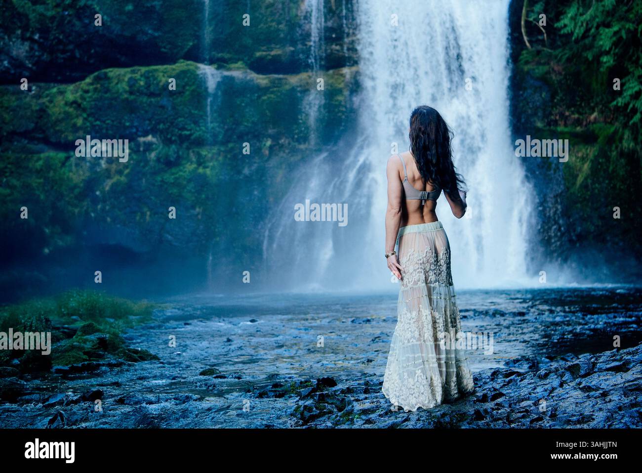 Frau in einem langen Rock steht an einem üppigen Wasserfall, umgeben von moosbedeckten Felsen. Silver Falls, Oregon, USA Stockfoto