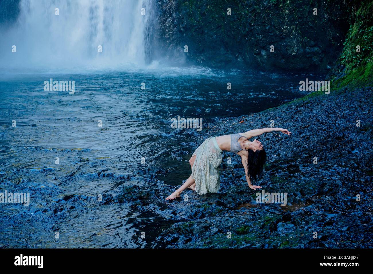 Frau in anmutigem Tanz posiert auf felsigem Gelände in der Nähe eines Wasserfalls und üppigem Grün. Silver Falls, Oregon, USA Stockfoto