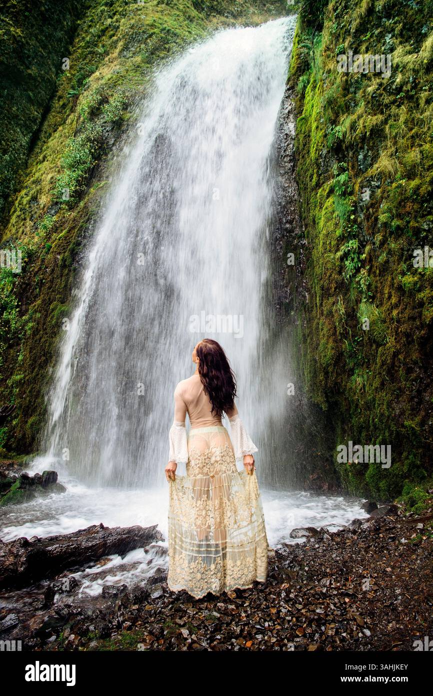 Frau in einem langen Rock steht mit Blick auf einen üppigen Wasserfall inmitten von leuchtend grünem Laub. Silver Falls, Oregon, USA Stockfoto