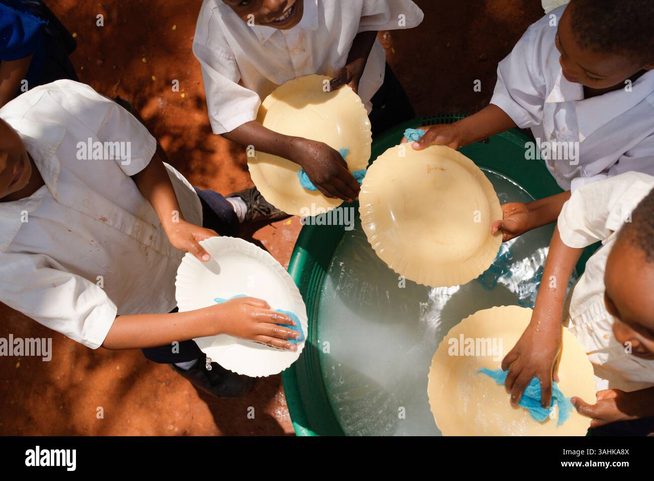 Kinder in weißen Hemden waschen Teller mit Schwämmen in einem Wasserbecken im Freien. Tansania, Afrika Stockfoto
