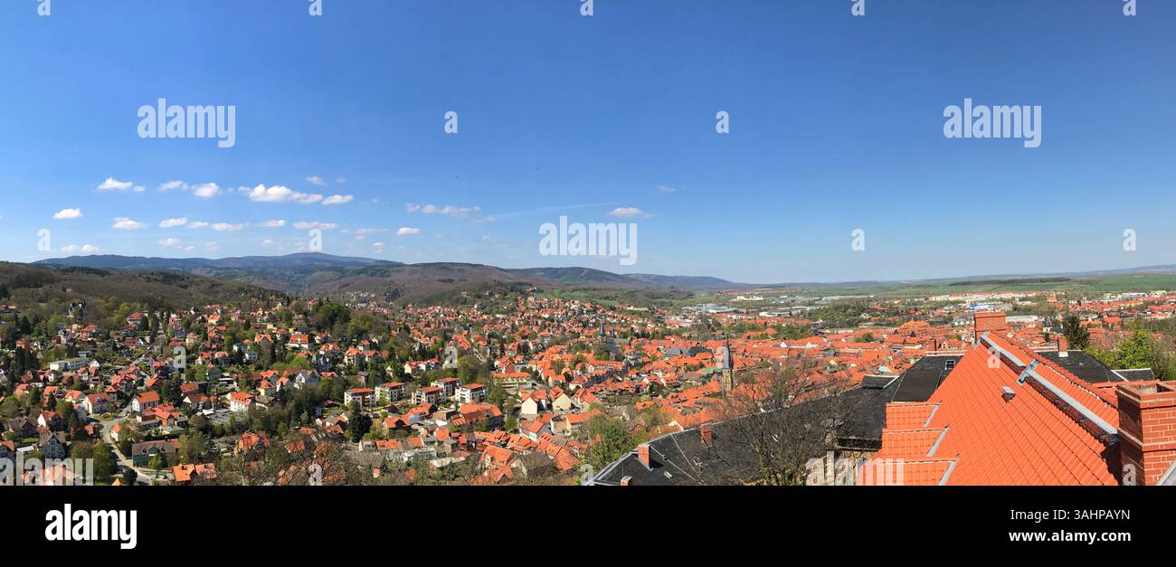 Ein malerisches Panorama, das eine Stadt mit roten Dächern, üppigem Grün und fernen Hügeln unter einem hellblauen Himmel zeigt, was ihre friedliche und malerische Umgebung hervorhebt Stockfoto