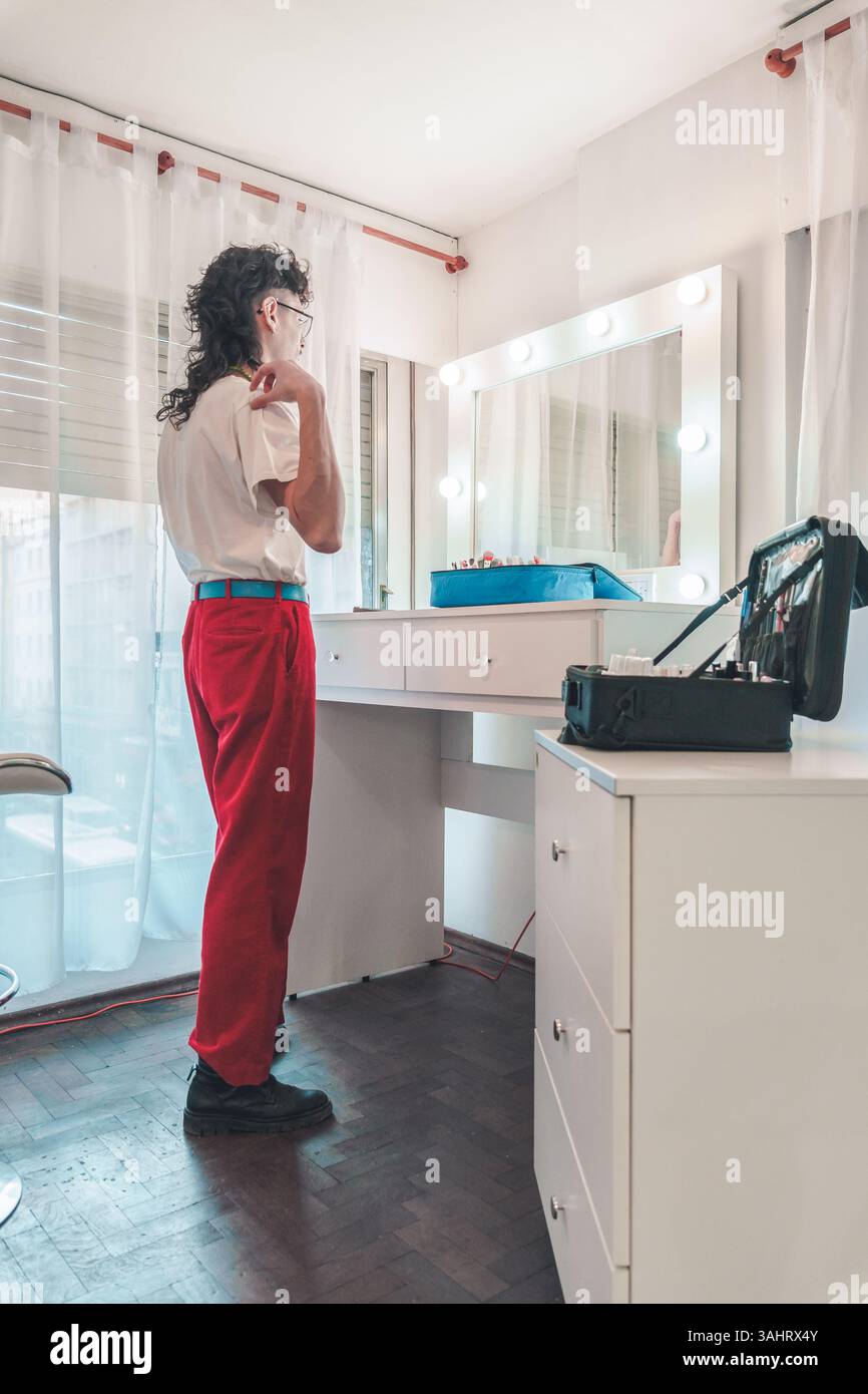 Eine nicht-binäre Person, die rote Hosen und ein weißes T-Shirt trägt, schminkt sich in einem hell beleuchteten Ankleidezimmer und hebt Themen der Selbstausdruckskraft hervor. Stockfoto