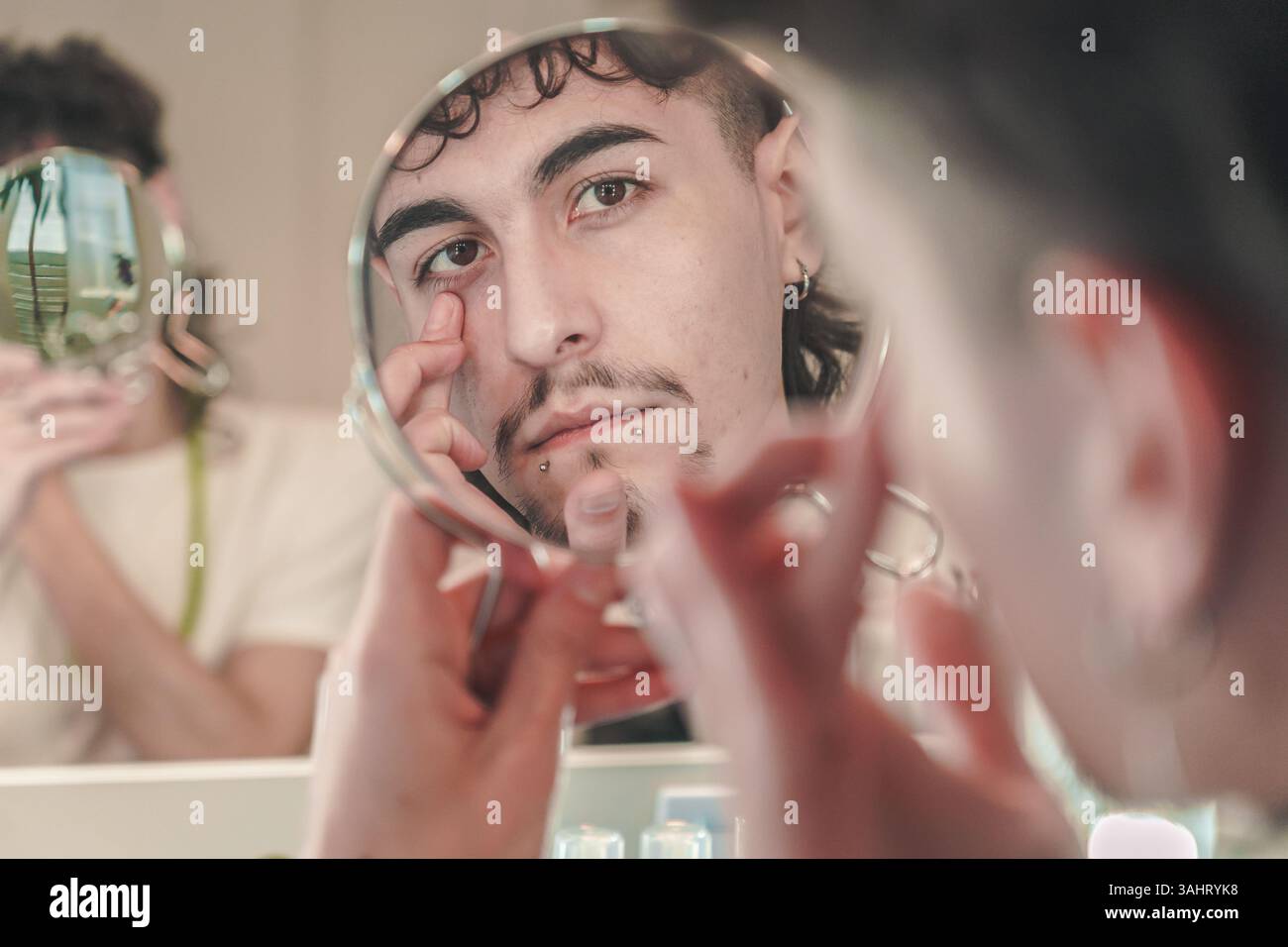 Ein junger Mann mit Piercings und gestylten Haaren untersucht sein Gesicht in einem kleinen Handspiegel und erkundet dabei Themen wie Selbstwahrnehmung, Identität und Vielfalt Stockfoto