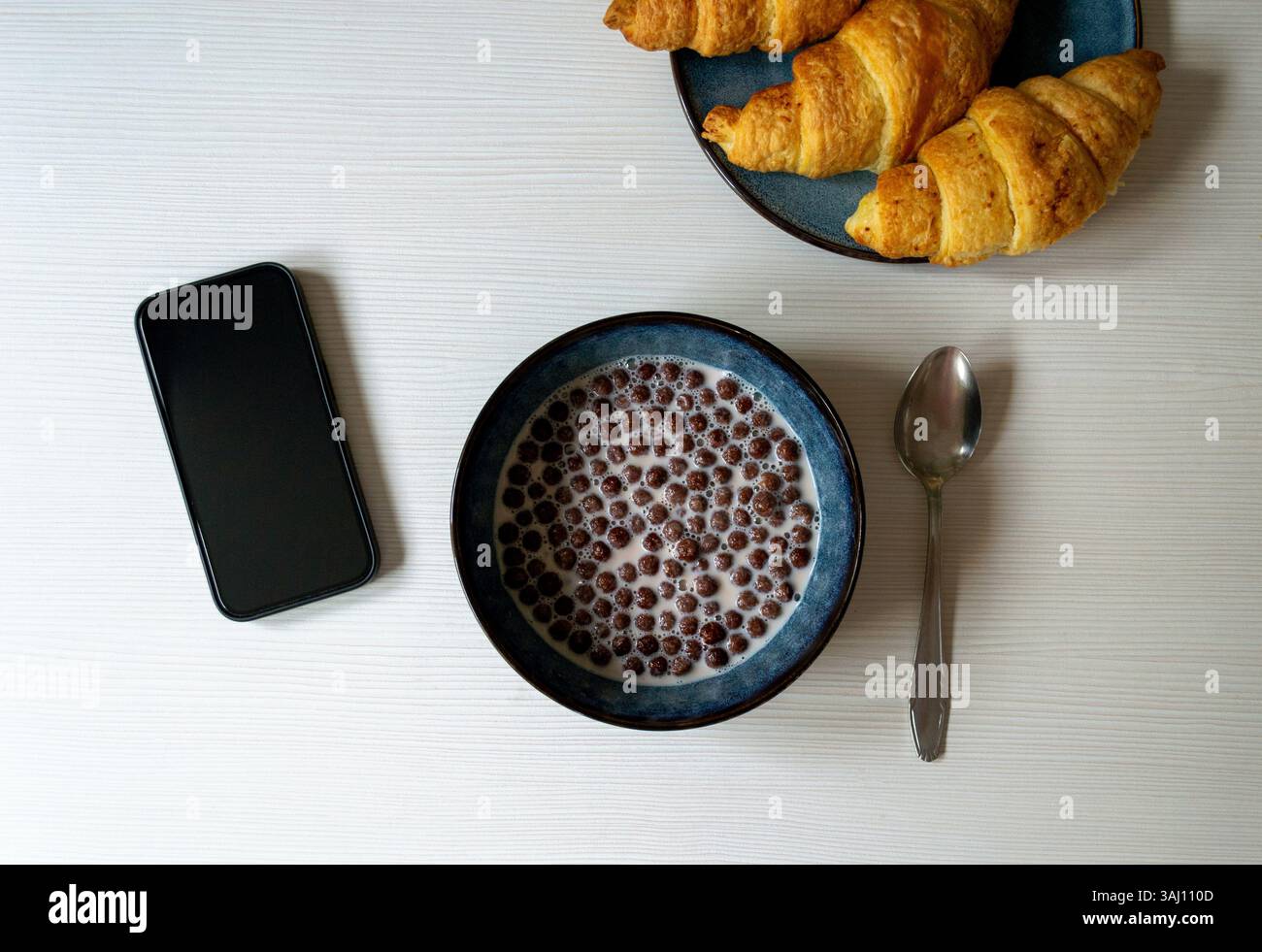 Frühstück: Müsli, Schokoladenkugeln mit Milch, Croissants und Smartphone auf dem Tisch, Flatlay Stockfoto