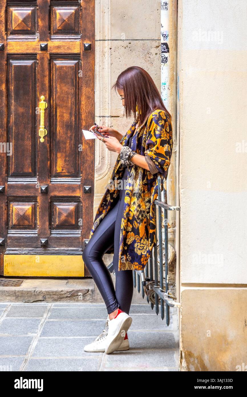 Junge, moderne, gut gekleidete asiatische Frau vor der Tür, die auf ihr Smartphone schaut - Paris, Frankreich. Stockfoto