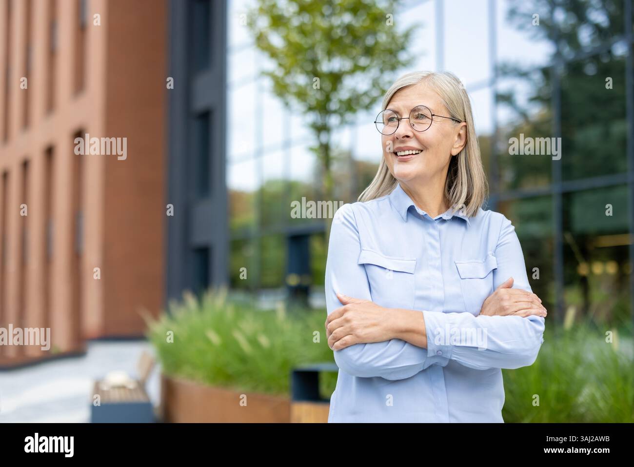 Eine lächelnde, reife Geschäftsfrau steht draußen mit überkreuzten Armen in der Nähe eines Bürogebäudes. Sie blickt auf etwas mit Optimismus. Stockfoto