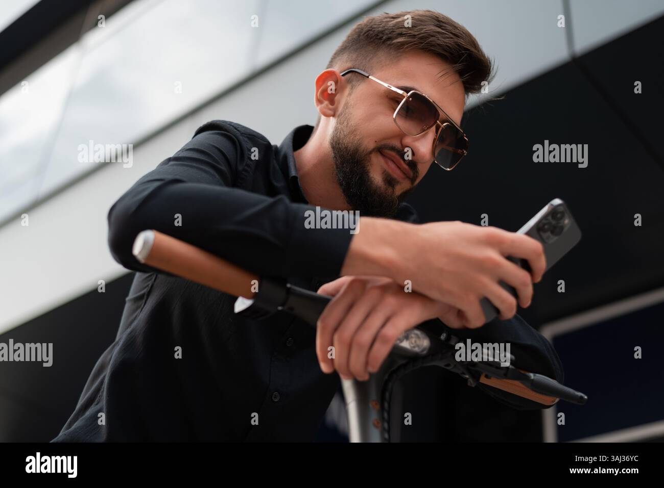 Ein junger Hipster-Mann mit einem Elektroroller, der vor einem modernen Geschäftsgebäude auf der Stadtstraße steht und ein Mobiltelefon benutzt. Stockfoto