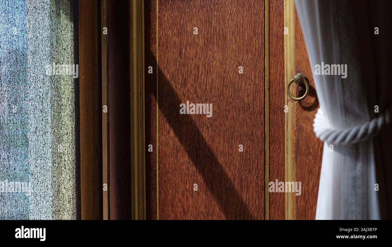Sanftes Sonnenlicht strömt auf eine Holztür mit Ringgriff und festgebundenem weißen Vorhang und offenbart eine ruhige, warme Innenatmosphäre. Stockfoto