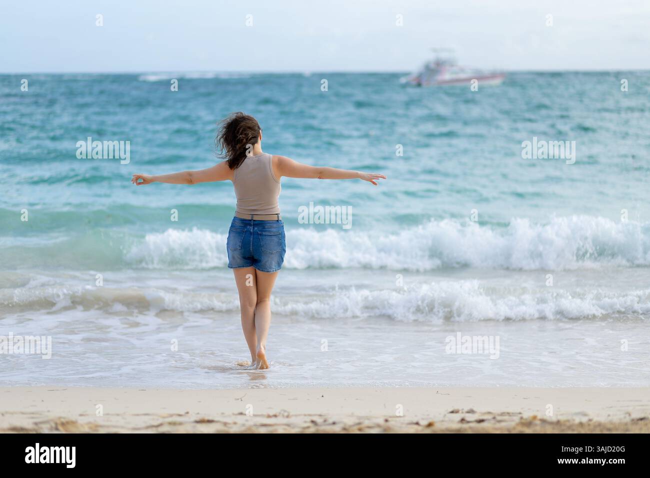 Eine Frau steht im flachen Wasser, die Arme gestreckt und die Meeresbrise umhüllt. Sanfte Wellen schlagen zu ihren Füßen, während ein Boot unter A über den Horizont fährt Stockfoto