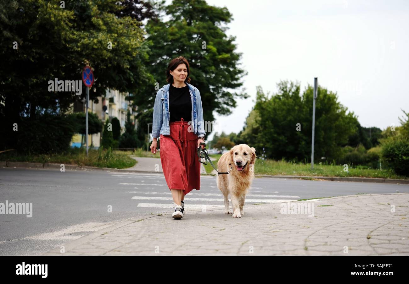 Eine Frau mit Einem Hund an der Leine überquert die Straße an Einem Querweg und geht durch Eine City Street Stockfoto