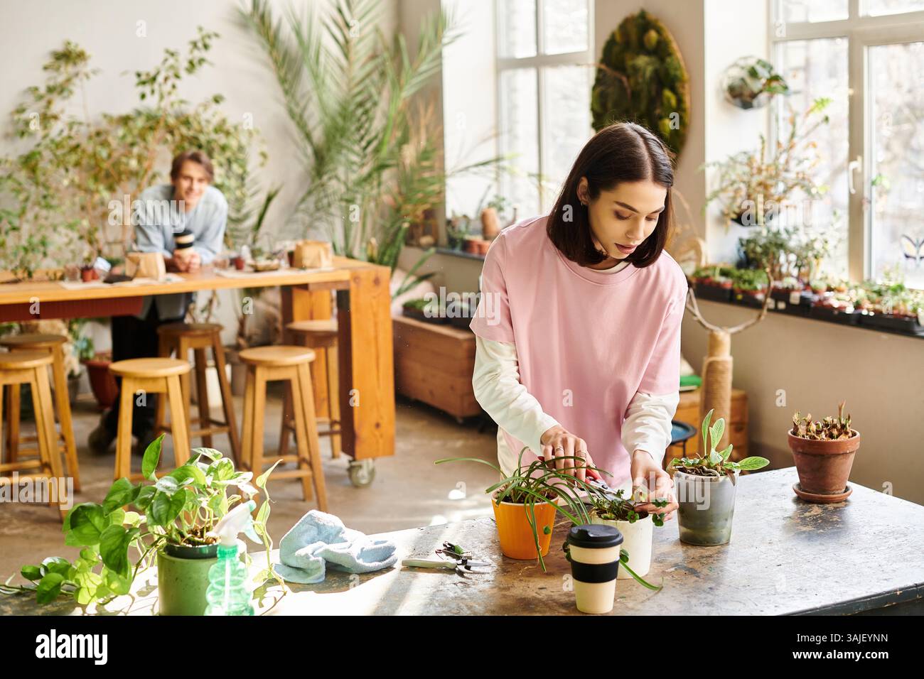 Eine junge Frau neigt dazu, Pflanzen zu Topfen, in Kreativität getaucht, während ein Mann aus der Ferne beobachtet. Stockfoto