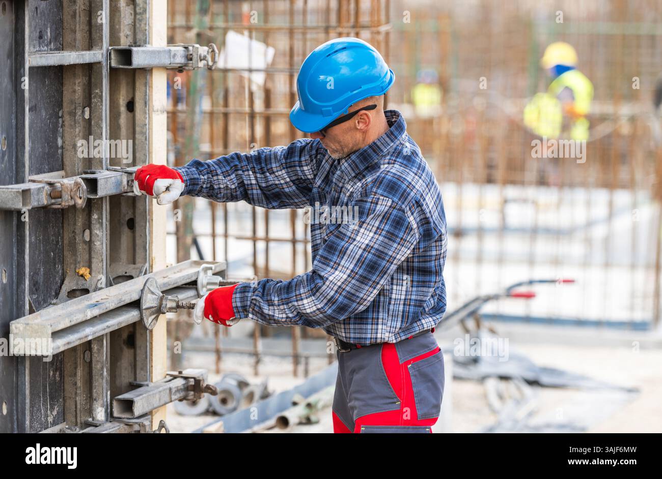 Ein Bauarbeiter in blauem Schutzhelm und kariertem Hemd passt Metallformen auf einer Baustelle an. Andere Mitarbeiter sind im Hintergrund sichtbar, engagiert Stockfoto