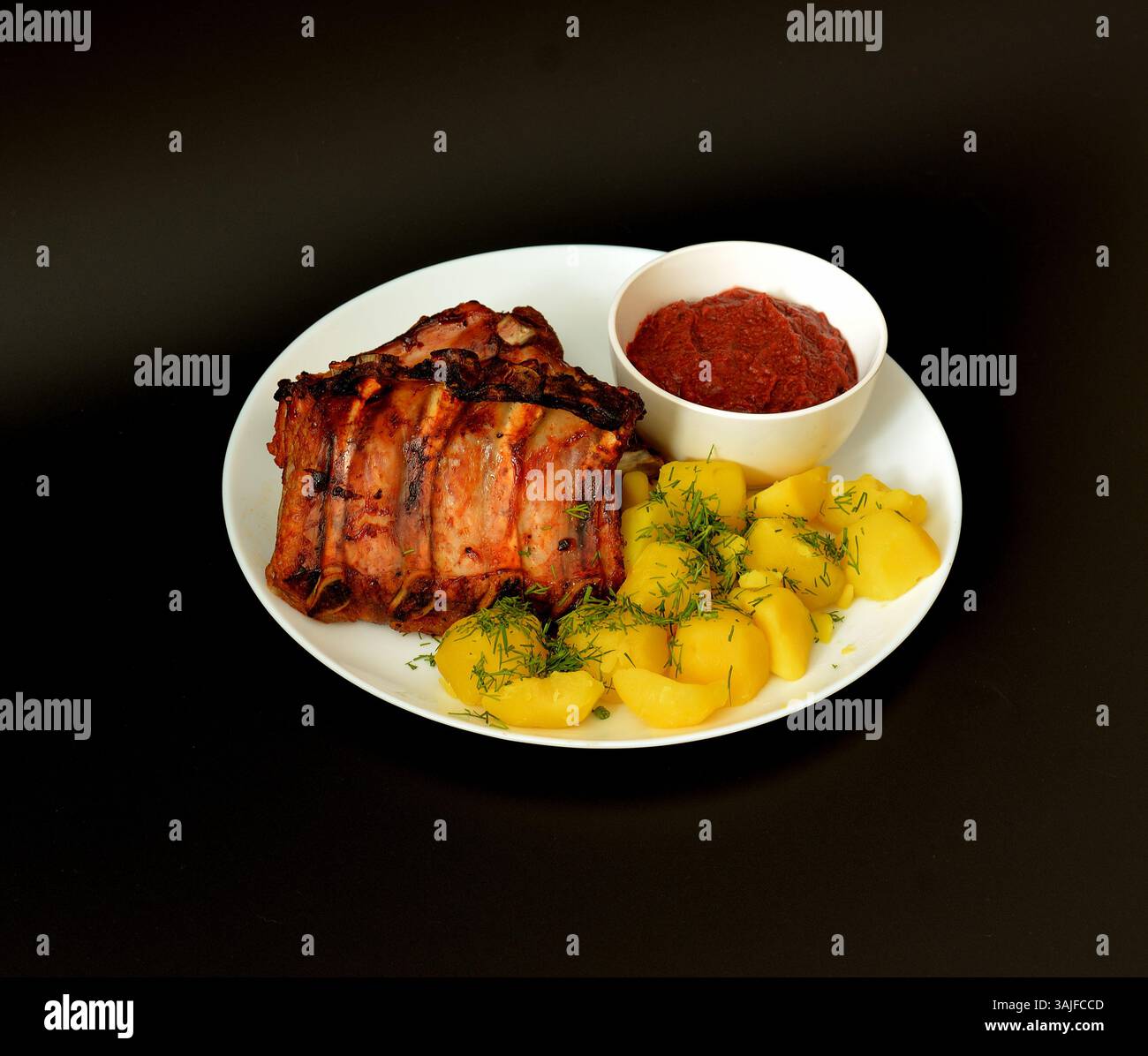 Gebackene Schweinerippchen mit Tomatenwürzsauce und jungen gekochten Kartoffeln auf einer großen weißen Keramikplatte auf schwarzem Hintergrund. Nahaufnahme. Stockfoto