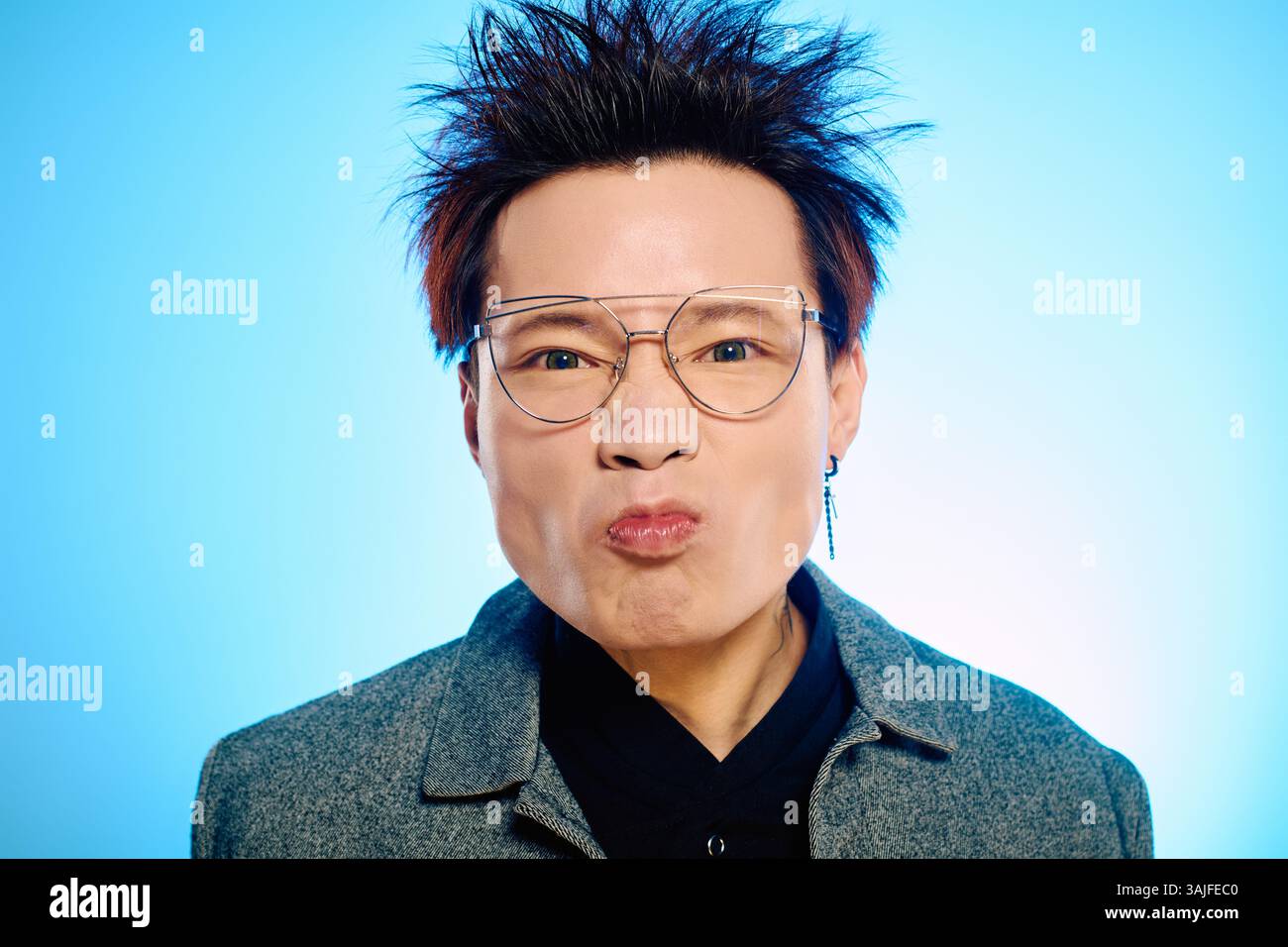 Der junge Mann zeigt einen verspielten Ausdruck mit auffälliger Frisur vor einem leuchtend blauen Hintergrund. Stockfoto