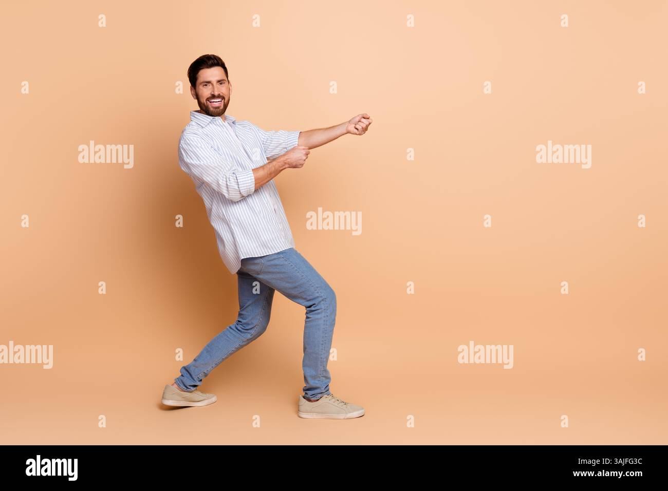 Ein lächelnder junger Mann posiert in einem stilvollen, legeren Outfit vor beigefarbenem Hintergrund mit einer verspielten Geste, an einem imaginären Seil zu ziehen. Stockfoto