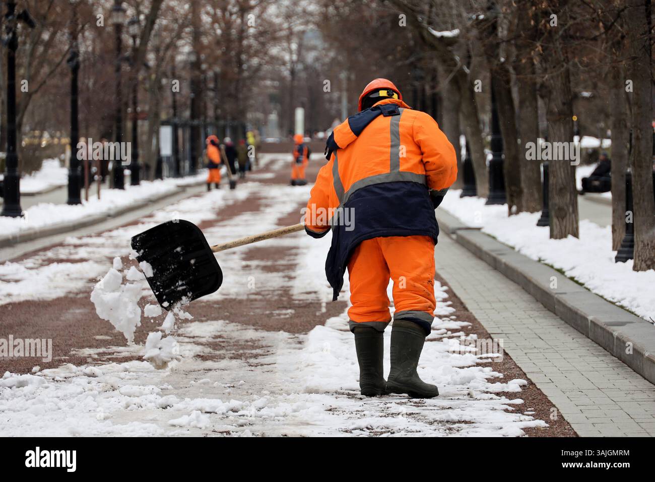 Kommunaler Arbeiter in Uniform mit Schaufel räumt Schnee auf dem Gehweg. Schneeräumung in Frühlingsstadt, Straßenreinigung im Park Stockfoto