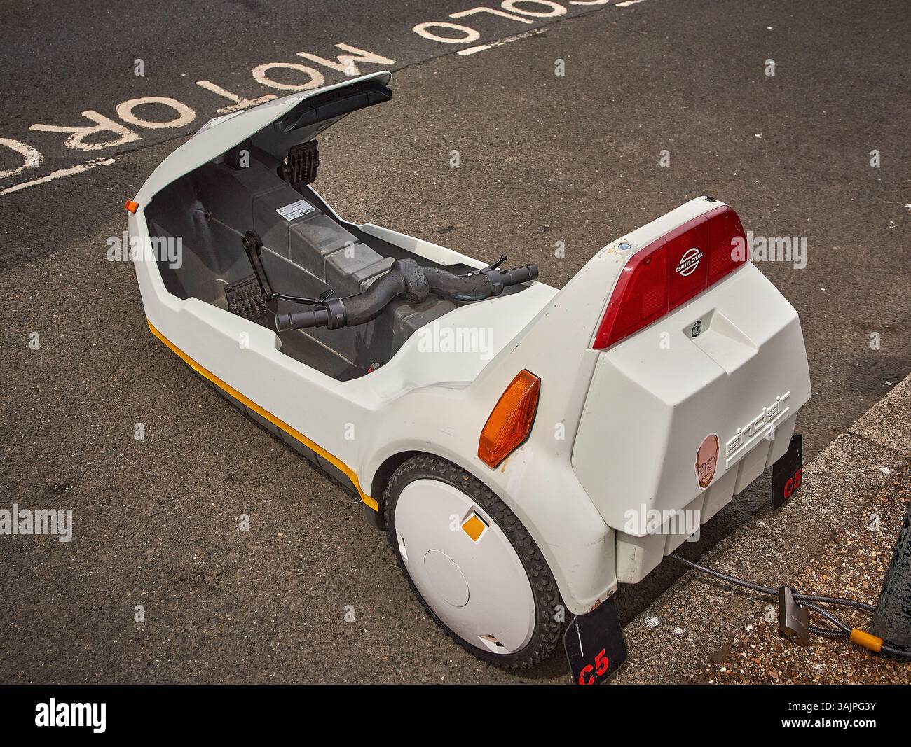 Hohe Blickwinkel-Rückseitenansicht eines weißen Sinclair C5 Elektro-Mikrofahrzeugs, das neben einem ausgewiesenen Motorradparkplatz abgesperrt ist. Stockfoto