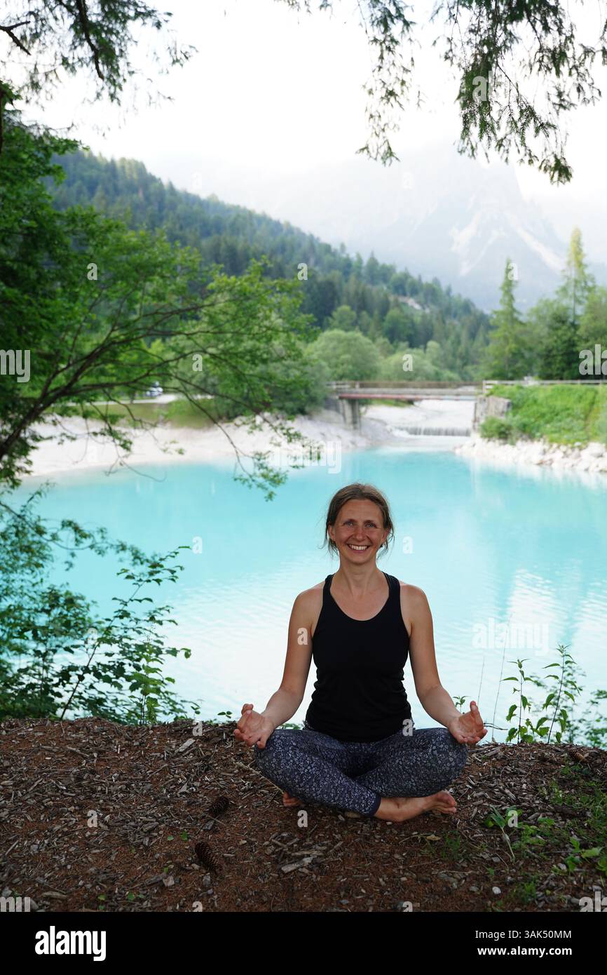 Frau, die friedlich im Freien meditiert, in einer ruhigen Natur neben einem ruhigen türkisfarbenen See, umgeben von üppigem Grün und Bergen. Sie sitzt Stockfoto