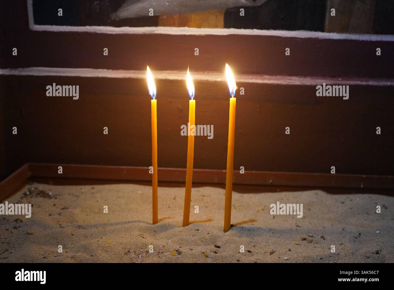 Drei schlanke, gelbe Kerzen stehen aufrecht auf einer sandigen Oberfläche in einer griechischen Kirche. Stockfoto