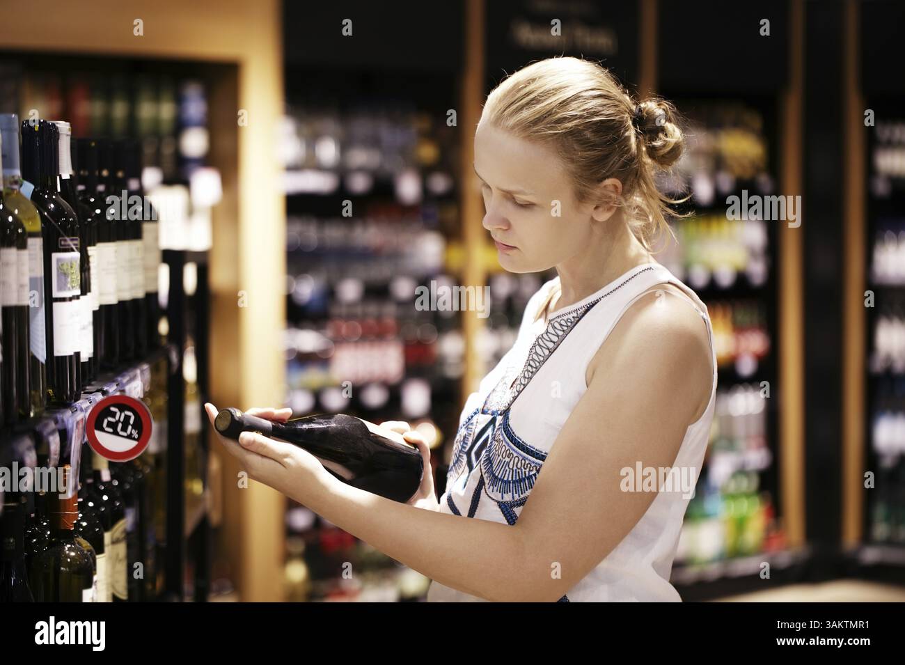 Frau, die Wein oder anderen Alkohol in einem Flaschengeschäft kauft, steht vor Regalen voller Flaschen und hält Flasche in der Hand und liest ins Buch Stockfoto