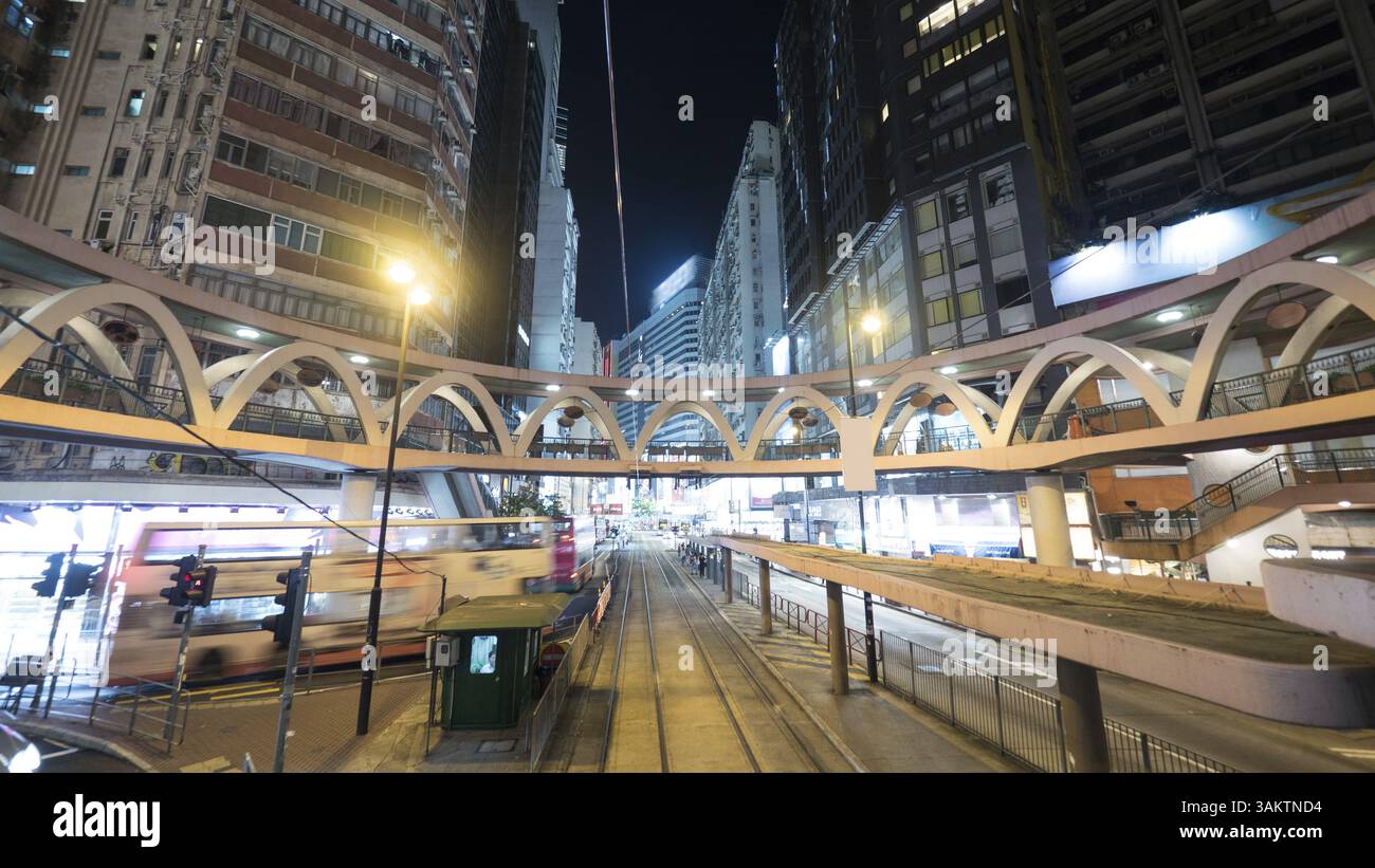 Nacht beleuchtet Hong Kong. Blick auf die Straßenbahnhaltestelle, die Fußgängerbrücke und den sich bewegenden Doppeldeckerbus zwischen Hochhäusern Stockfoto