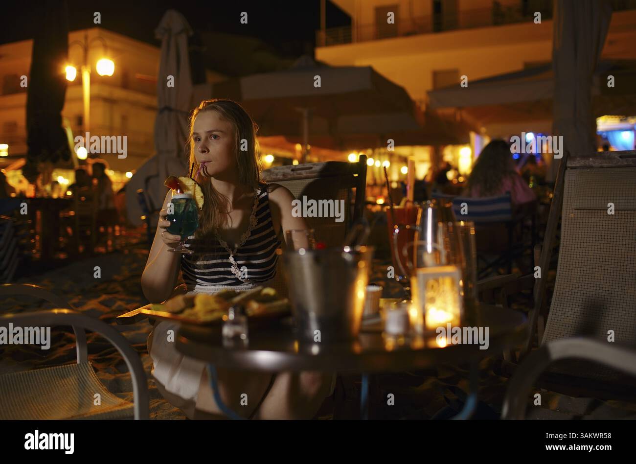 Junge Frau mit einem Drink in einem Pub oder Restaurant an einem Tisch allein im Freien durch elektrische Lampen bei einem Drink beleuchtete Sitzen Stockfoto