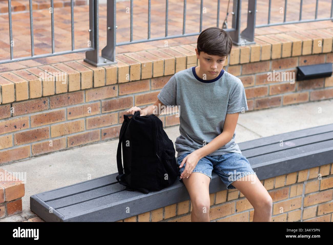 Sitzt auf der Bank, der Junge hält den Rucksack und sieht nachdenklich in der Schule aus Stockfoto