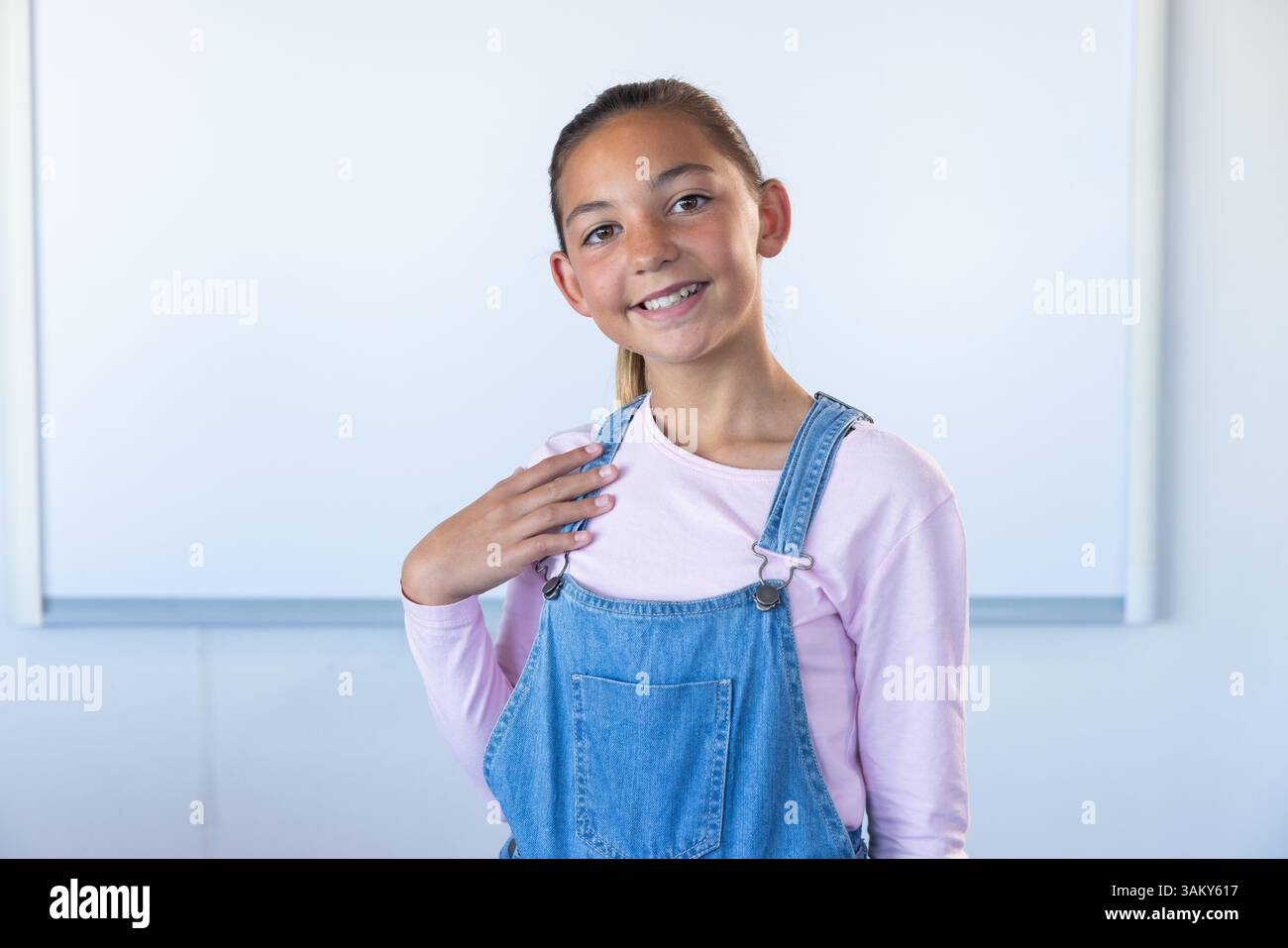 Lächelndes Mädchen in der Schulklasse, trägt Denim-Overall und rosa Hemd Stockfoto