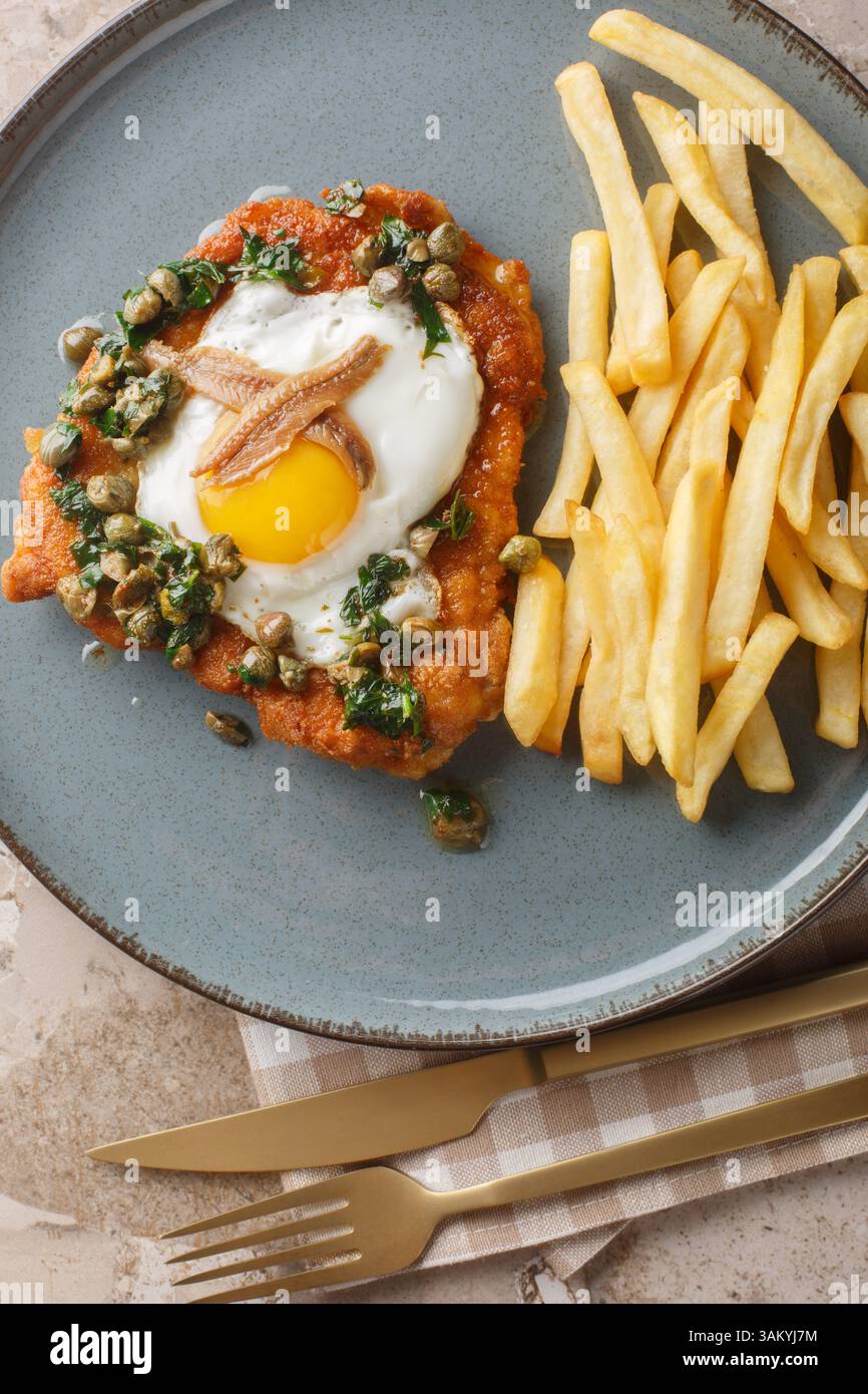 Holsteinschnitzel mit Sardellenfilet, Spiegelei und Kapern-Petersiliensauce darauf serviert mit Pommes Frites in Nahaufnahme auf einem Teller auf einem Tisch. Vertikal oben Stockfoto