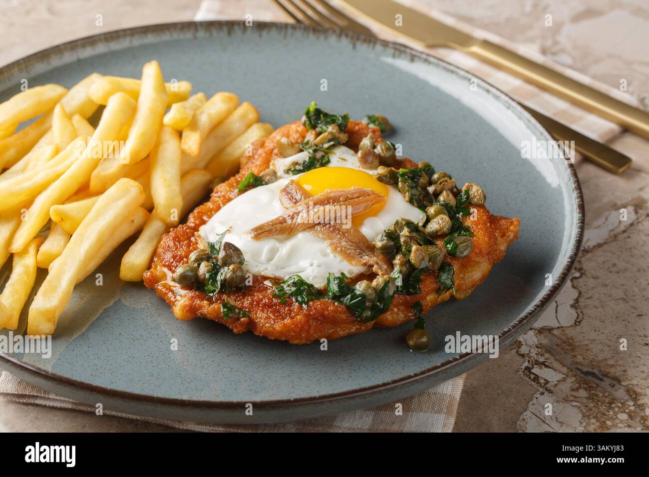 Köstliches paniertes Schnitzel mit Sardellen, Spiegelei und Kapern Petersiliensauce, serviert mit pommes Frites in Nahaufnahme auf einem Teller auf dem Tisch. Horiz Stockfoto