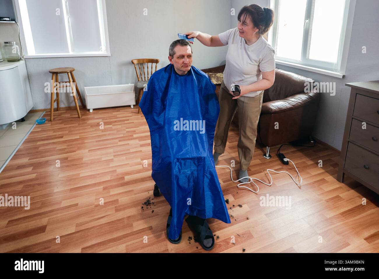 Das Familienmitglied schneidet die Haare reifer Erwachsener mit einem Haarschneider in Innenräumen, mit blauem cape um die Schultern und Holzboden darunter. Stockfoto