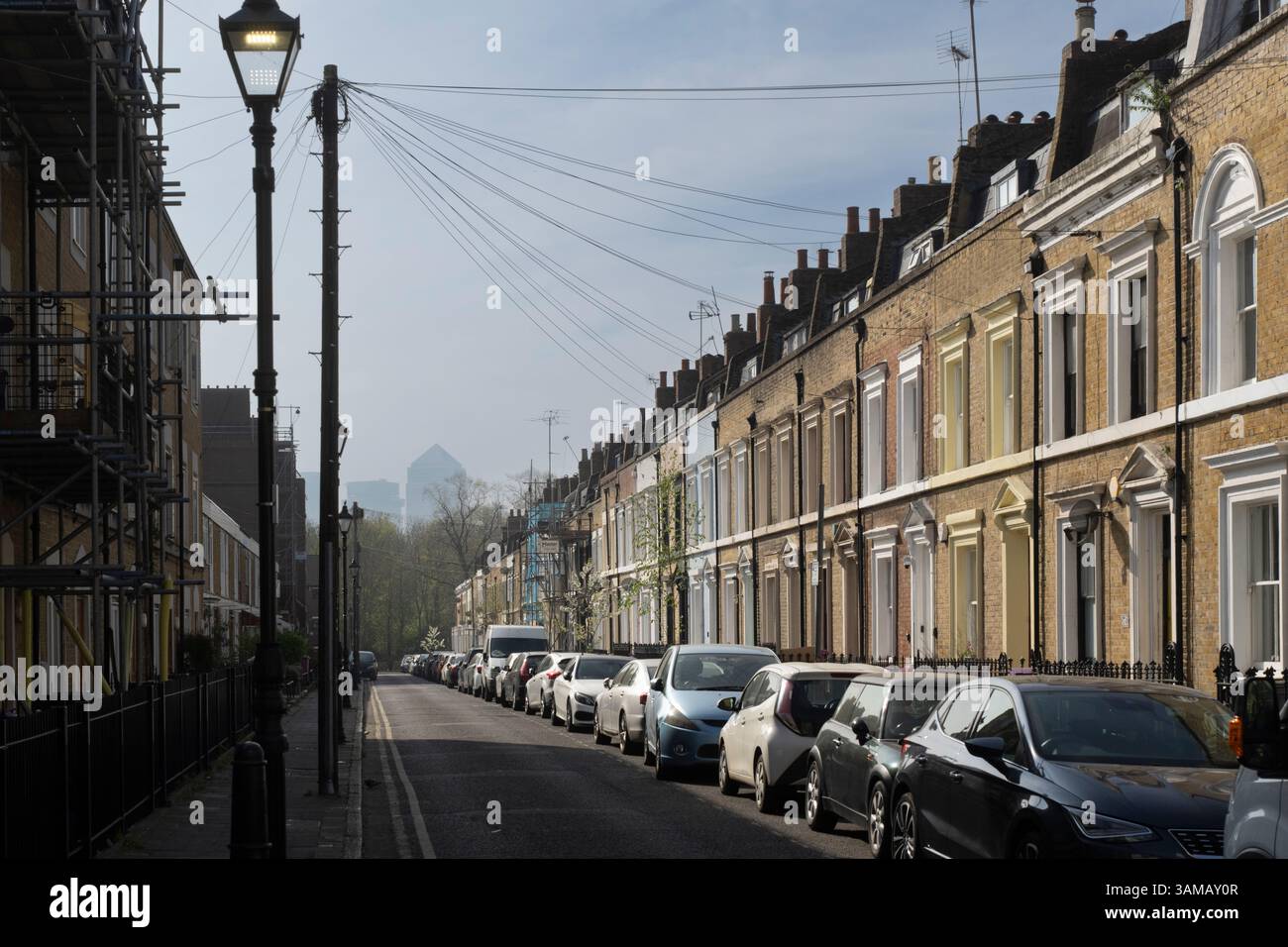 Autos säumen eine Straße mit Reihenhäusern, Brokesley Street, Mile End, London. Die Skyline von Canary Wharf ist in der Ferne sichtbar. Stockfoto